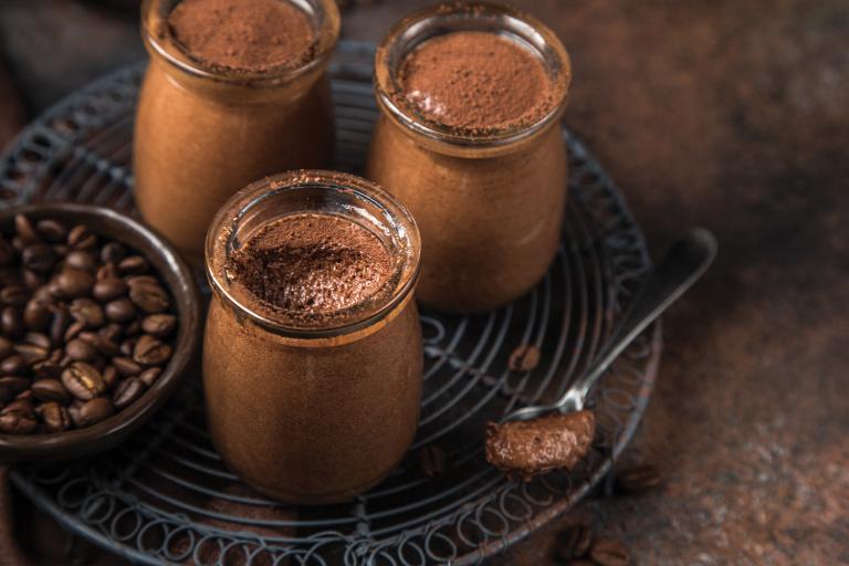 Chocolate mousse in clear glass jars, next to a small dish of coffee beans on a wire metal trivet.